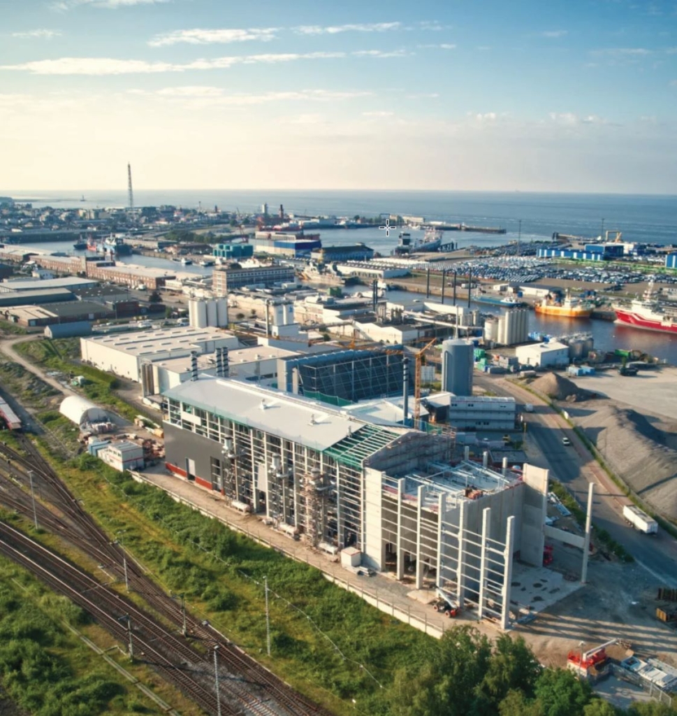 Luftaufnahme eines Industriehafengebiets mit groÃŸen LagerhÃ¤usern, FabrikgebÃ¤uden, KrÃ¤nen und Schiffen in KÃ¼stennÃ¤he.