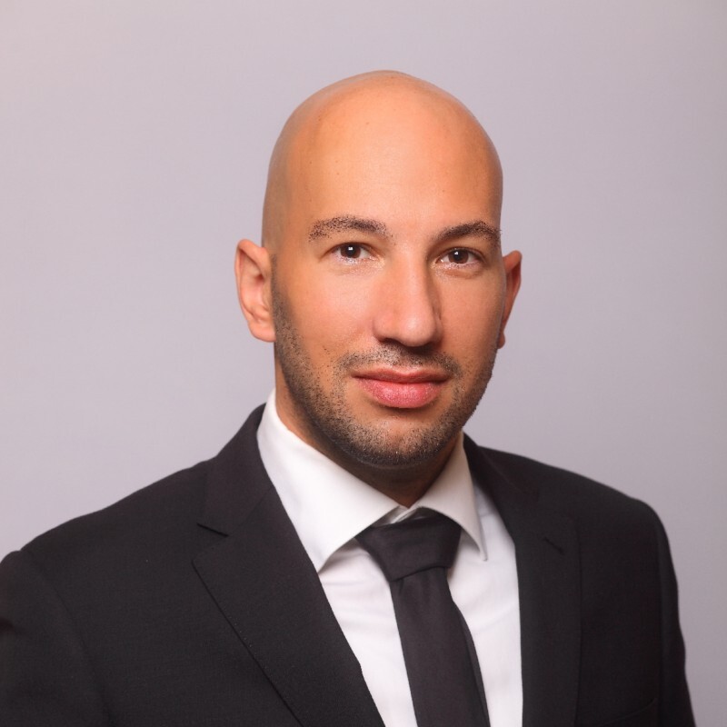 Ein glatzkÃ¶pfiger Mann in Anzug und Krawatte, der selbstbewusst und mit neutraler Miene dasteht.