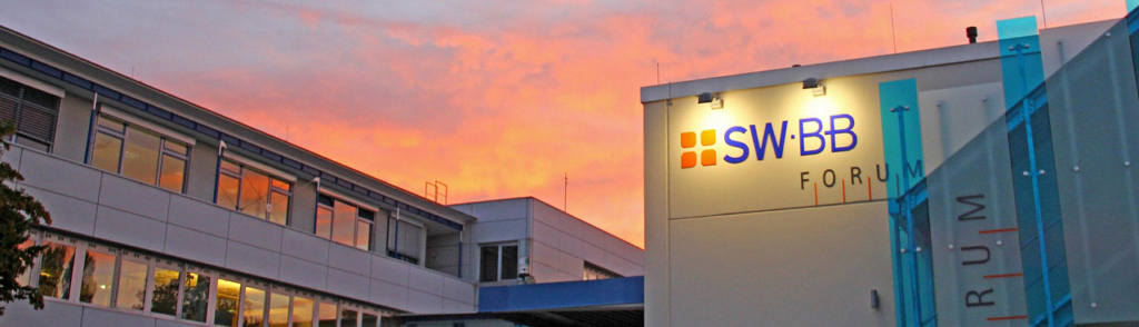 Modernes GebÃ¤ude mit beleuchtetem SWBB-Forum-Logo an der Fassade, unter einem farbenfrohen Sonnenuntergangshimmel mit orangefarbenen und rosafarbenen TÃ¶nen.