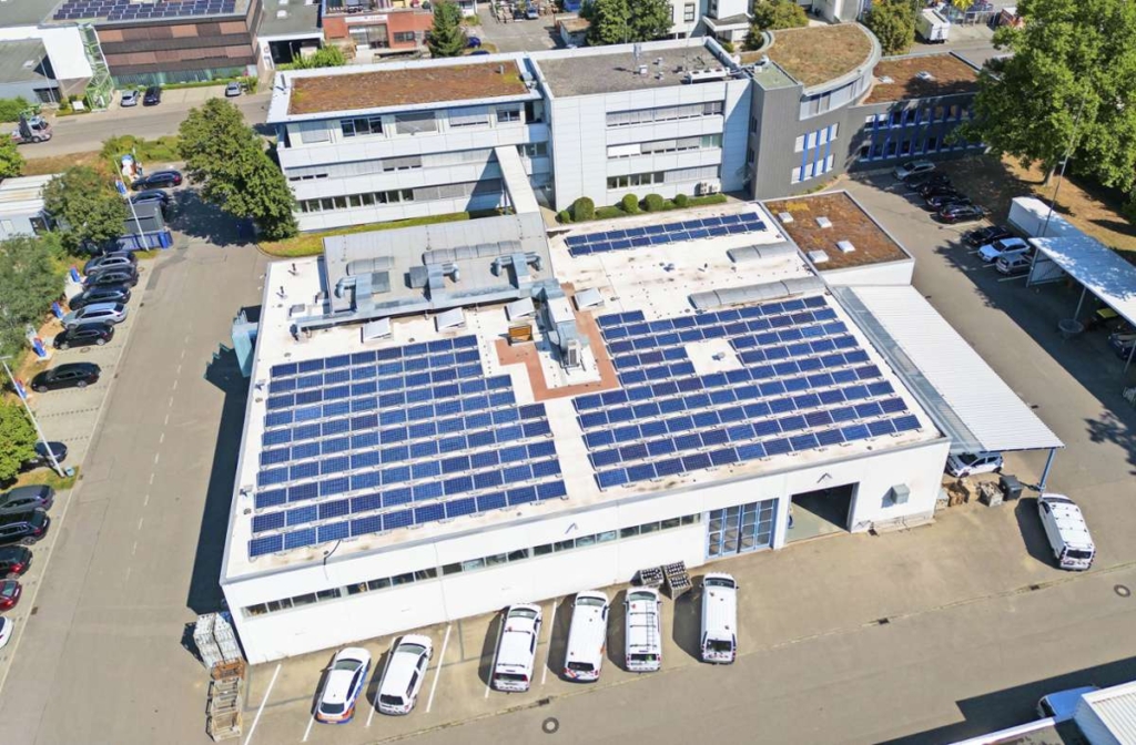 Luftaufnahme eines groÃŸen IndustriegebÃ¤udes mit zahlreichen Sonnenkollektoren auf dem Flachdach. Das GebÃ¤ude ist umgeben von geparkten Autos und anderen GebÃ¤uden mit begrÃ¼nten DÃ¤chern und BÃ¤umen in der NÃ¤he.