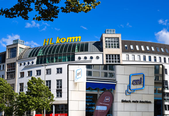 HL-Komm-GebÃ¤ude mit gewÃ¶lbtem Glasdach und Beschilderung, mit zusÃ¤tzlicher Ladenfront mit dem Branding â€žen! Einfach. mehr Anschluss.â€œ unter strahlend blauem Himmel.