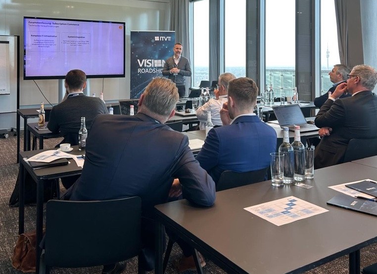 Ein professioneller Workshop oder eine PrÃ¤sentation in einem modernen Konferenzraum. Mehrere Teilnehmer sitzen an Tischen und blicken auf einen Referenten, der neben einem groÃŸen Bildschirm steht, auf dem eine Folie gezeigt wird. Neben dem Bildschirm befindet sich ein vertikales ITVT-Banner mit dem Titel â€žVISION ROADSHOWâ€. Auf den Tischen stehen Wasserflaschen, NotizbÃ¼cher, Laptops und gedruckte Unterlagen. Durch die groÃŸen, vom Boden bis zur Decke reichenden Fenster im Hintergrund hat man einen Blick auf die Stadt.