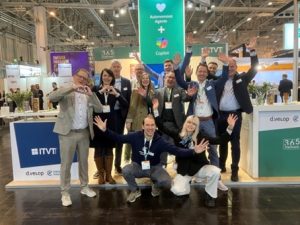 Eine Gruppe von Menschen posiert voller Energie für ein Gruppenfoto am Messestand von ITVT. Der Stand ist mit auffälligen Schildern ausgestattet, darunter Banner für ITVT, d.velop und 365 Stadtwerk. Ein großes grünes Display im Hintergrund wirbt für „Autonomous Agents + Copilotâ€. Die Szene spielt in einer belebten Messehalle, in der überall Lichter, Stände und Besucher zu sehen sind.